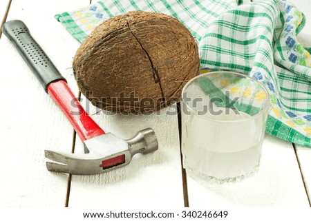 Cracked coconut, coconut water in a glass, hammer and napkin on a white wooden table. Selective focus