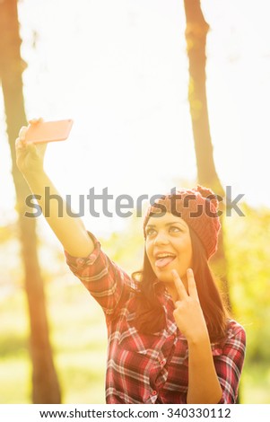 Modern young millennial woman in red plaid shirt and beanie hat taking a selfie on smartphone. Girl with tongue piercing posing taking self portrait in park in autumn. Back light, vibrant colors.