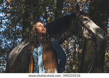 Pretty blonde girl standing by her grey horse in the countryside