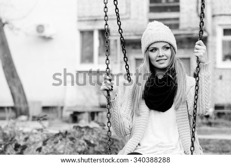 Close up outside portrait of beautiful smiling blonde girl sitting on a swing, wearing knitwear. Urban city scene. 