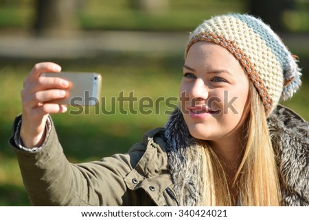 Cute, young girl with wool cap taking selfie by cell phone outdoors in a sunny autumn day