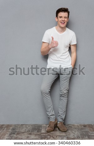 Full length portrait of cheerful handsome inspired confident young man in white t-shirt and gray pants showing thumbs up and winking on gray background