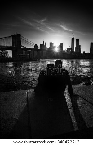 A couple in love - woman and man casting long shadows sitting on a pavement watching a sunset over the Hudson river, the Brooklyn bridge and Manhattan skyscraper skyline; New York City, United States