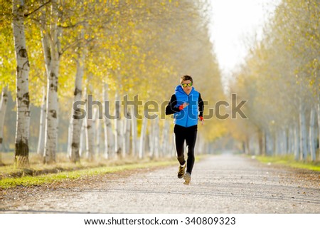 front view young sport man running outdoors in off road trail track with trees in Autumn sunlight wearing jogging vest and sunglasses in fitness, countryside training and healthy lifestyle concept