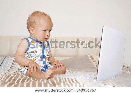 Adorable blond toddler boy sitting on the bed and looking at laptop at home, indoors. Child with tablet computer.