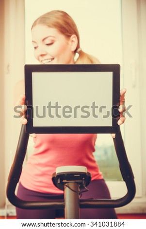 Active young woman working out on exercise bike stationary bicycle holding pc tablet computer with blank screen copyspace. Sporty girl training at home. Fitness and weight loss advertisement concept.