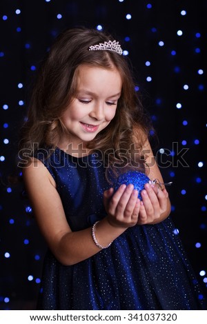Happy pretty  child girl is holding blue Christmas tree ball in hands over background scene with lights, New years
