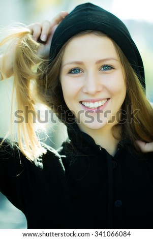 Portrait closeup of smiling pretty blond girl with long hair wearing black hood taking hair in hands posing on streetscape background, vertical picture