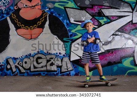 Confident small skateboarder posing with crossed arms in front of colorful graffiti painted on a wall with his skateboard on the ground beside him