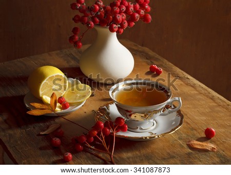 Cup with hot tea, a lemon and a jug with ash berries on a rough wooden surface. Beautiful autumn still life. 