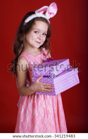 Cute little happy girl is wearing pink dress and bunny ears on red background is holding gift boxes