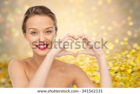 beauty, people, love, valentines day and make up concept - smiling young woman with pink lipstick on lips showing heart shape hand sign over golden glitter or holidays lights background