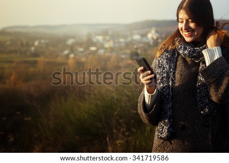 gorgeous stylish beautiful happy brunette girl looking at the telephone and smiling on a background of sunny autumn nature