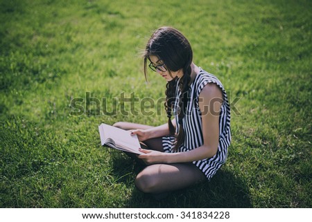 Beautiful Dark-haired Young Woman Sits On Grass And Reading Book, Against Summer Green Park.