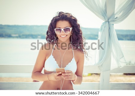 Closeup young beautiful happy excited smiling woman with mobile cell  smart phone  listening to music outdoors on sun-bed, sea lake background looking at you. Positive facial expression human emotion