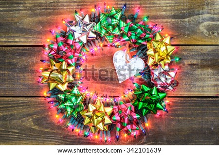 Christmas wreath with colorful holiday lights, silver tin heart and pink, gold and green bows on antique rustic wood background