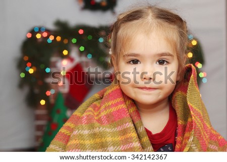 Happy little girl covering herself up in blanket over blurred Christmas decorated fireplace on background, winter holiday family concept