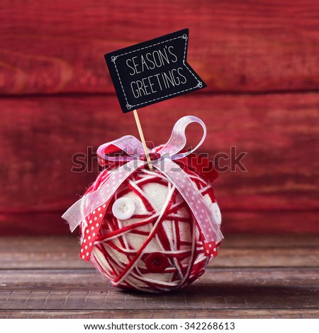 a handmade christmas ball, made with different ribbons and buttons, topped with a flag-shaped signboard with the text seasons greetings, placed on a rustic wooden surface