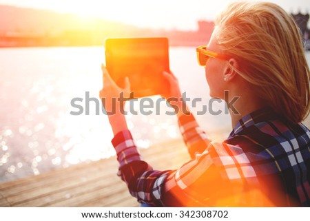 Young stylish hipster girl photographing on digital tablet beautiful sea landscape in summer evening, woman taking photo with touch pad camera while sitting on a river jetty during recreation time 