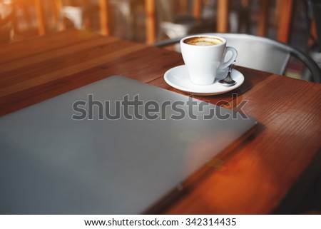 Closed laptop computer and cup of hot cappuccino lying on a wooden table in contemporary coffee shop interior, portable net-book with space for your brand, electronic business remote job via internet 