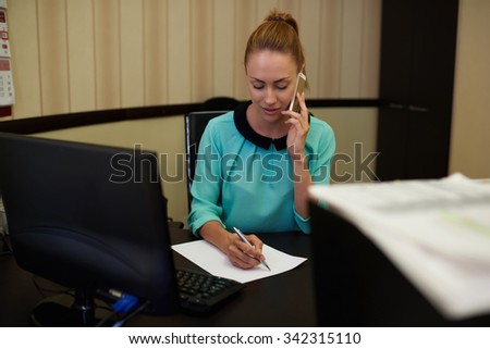 Young attractive businesswoman sitting at workplace and signing papers in comfy office, young female entrepreneur converting on mobile phone next to table pc computer focuses on professional process 