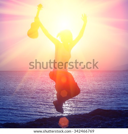 Silhouette of young girl jumping with hands up, with ukulele, on the beach at the sunset