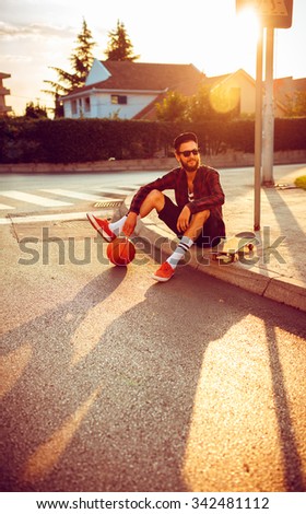 Young stylish man in sunglasses with a basketball and skateboard sitting on a city street at sunset light
