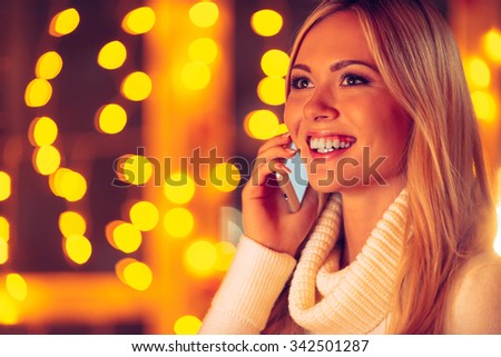 Staying connected. Beautiful young smiling woman in white sweater talking on the mobile phone with defocused Christmas lights in the background 