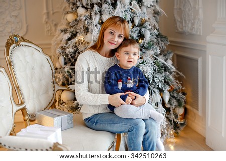 Mother in a white sweater smiles and holds a charming baby boy in blue sweater against the backdrop of Christmas trees in the interior of the house