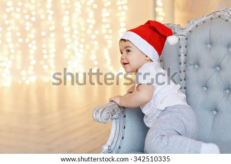 Small charming baby boy in red Santa hats sitting on a chair against a background of Christmas lights and smiling, in the interiors of the house
