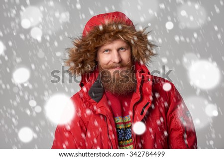 Closeup portrait of bearded man in red winter jacket with hood on, over snow background