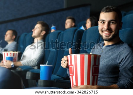 Movie mania. Young handsome bearded man showing thumbs up sitting in the cinema holding his popcorn 