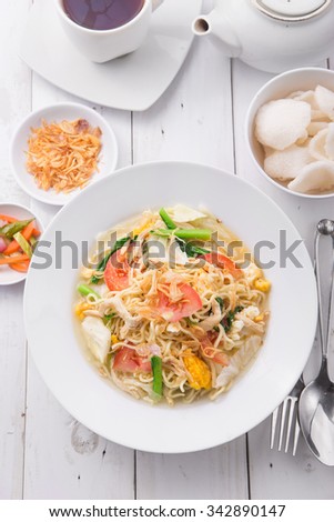 A bowl of noodles with eggs, chicken, tomato, green vegetables served with shrimp crackers, fried shallot, cayenne on rustic white wooden background