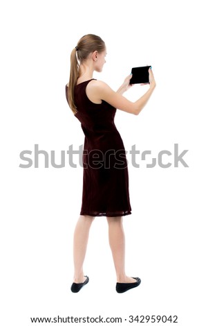 back view of standing young beautiful girl with tablet computer in hands of. girl  watching. Isolated over white background. The blonde burgundy dress holding to a horizontal smartphone