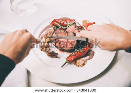 Person cuts beef steak with grilled vegetables served on white plate. Toned picture