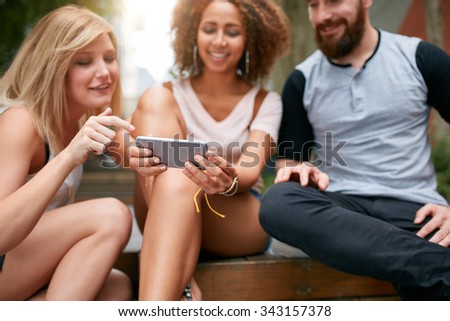 Close up shot of young adults looking something interesting on cellphone. Young friends using mobile phone while sitting together outdoors. Focus on smart phone in woman's hands. Royalty-Free Stock Photo #343157378