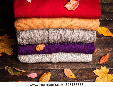 Pile of knitted winter clothes on wooden background covered with autumn leaves, knitwear, space for text. Stack of knitted sweaters and cardigans.