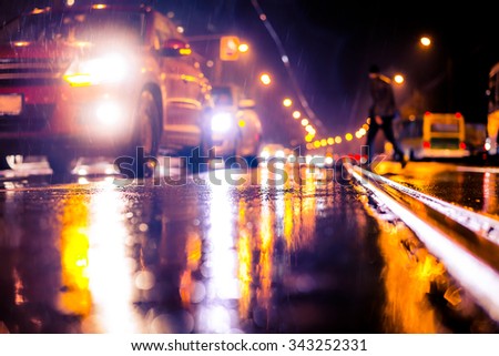 Rainy night in the big city, stream of cars traveling along the avenue and pedestrians on the road. View from the tram rail level, image in the soft orange-purple toning
