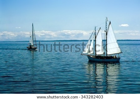 Sailboats off the Maine coast in placid water with blue sky and puffy clouds, a sunny day with 2 boats reflected in water 