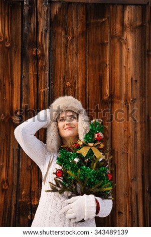 Christmas in the country style. Relaxed and dreamy young woman in white knitted sweater and furry hat holding Christmas tree in the front of rustic wood wall
