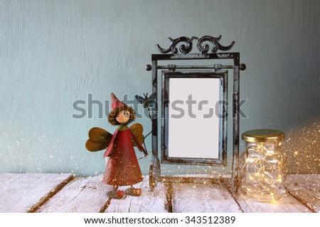 low key image of old victorian steel blue blank frame, mason jar with fairy lights and cute fairy on wooden table. vintage filtered image with glitter overlay