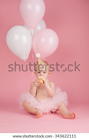 A portrait of a one year old baby girl wearing a pink tutu, string of pearls and birthday hat. She is sitting and eating a cupcake on a pink background.