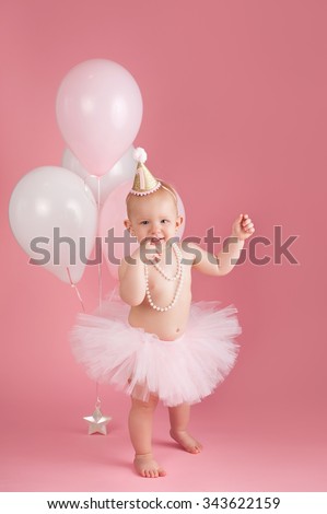 A portrait of a smiling one year old baby girl wearing a pink tutu, string of pearls and birthday hat. She is standing on a pink, background with a happy expression on her face.