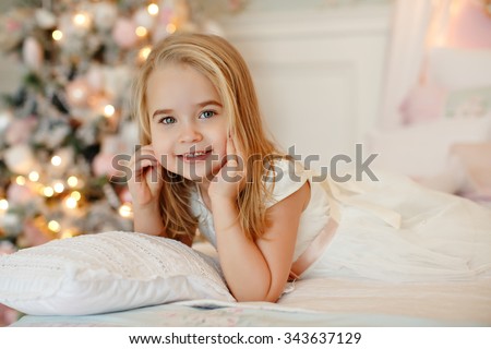 Very nice charming little girl blonde in white dress lying on the bed and looking at the picture on the background of smiling Christmas trees in bright interior of the house