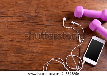 White cellphone with headphones and pink dumb bells on varnished wooden background
