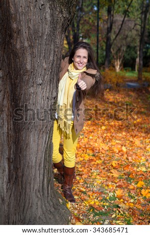 Happy woman peeking out from behind tree in park in autumn and showing thumb up.