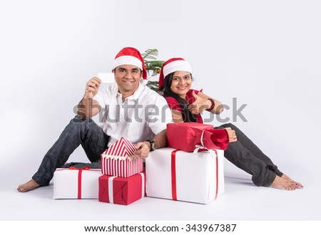 young indian couple sitting with with christmas gifts showing card, asian couple and christmas gift, with santa hat, white background, india and christmas celebration, christmas gift and indian couple