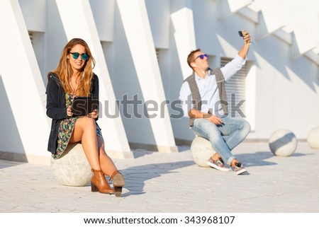 Sitting hipster couple using technology devices on white urban background - Tech concept with smiling girl holding tablet and listening music while guy take selfie portrait photo with smartphone