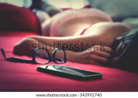 Hipster girl lying in the bed with her cellphone and eyeglasses.