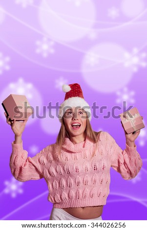 A portrait of beautiful delighted blonde lady wearing red Santa Claus hat and holding gifts in both hands on bright purple background